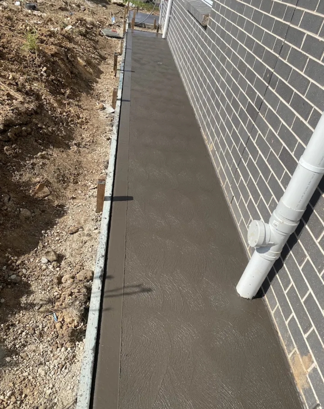 Concrete side path poured along a dark brick wall, edge formwork still in place